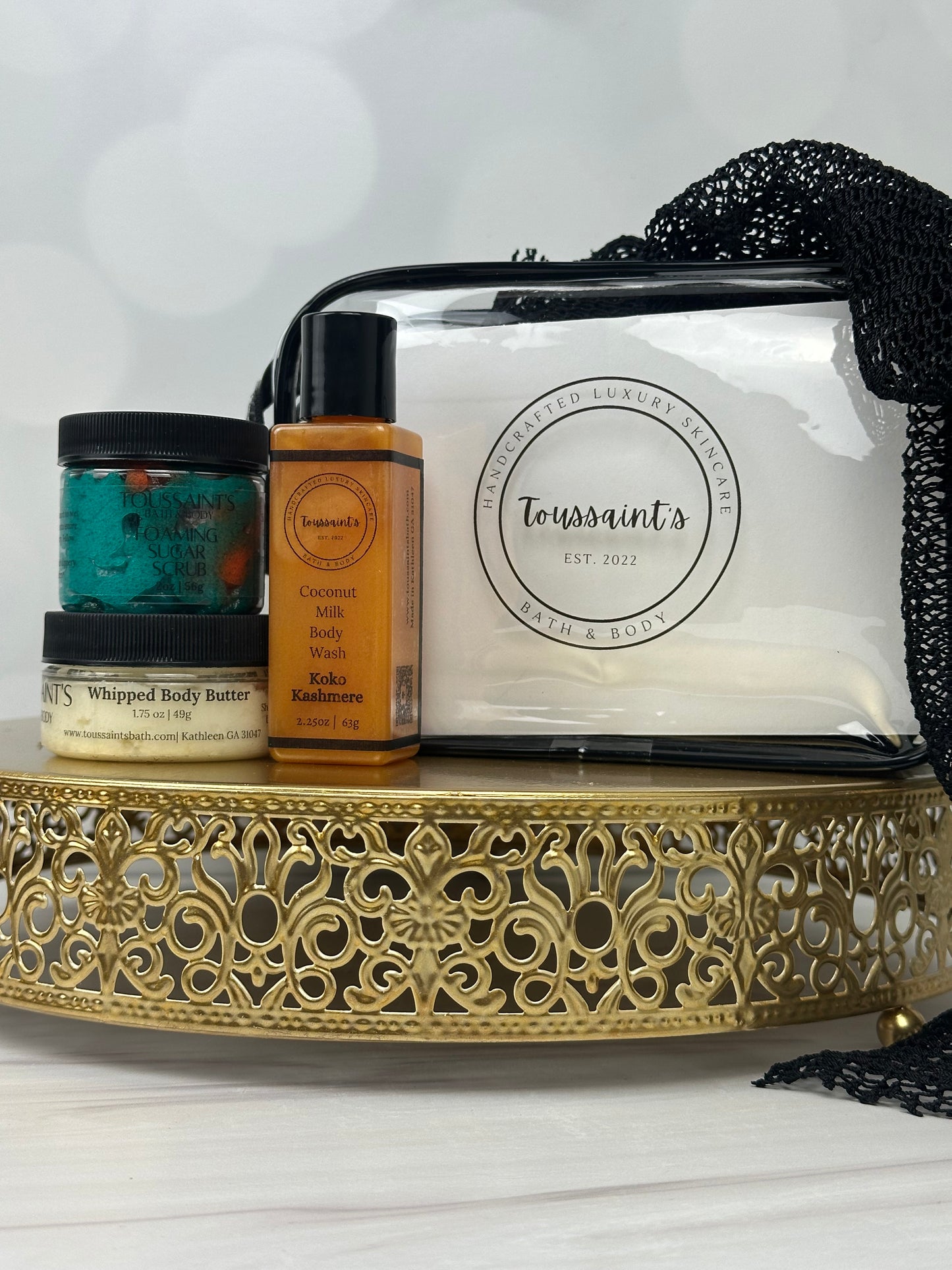 A curated gift set of wellness and beauty products arranged on a decorative gold tray with black lace. The set includes a lclear jar labeled whipped body butter, a small amber bottle, a teal jar, and a clear vinyl zipper bag, set against a white background.